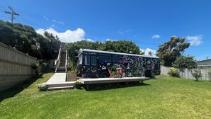 A bus with a colorful mural parked in a backyard with a wooden deck and stairs leading up to it. - Luxury Beach Bus (Whanganui)
