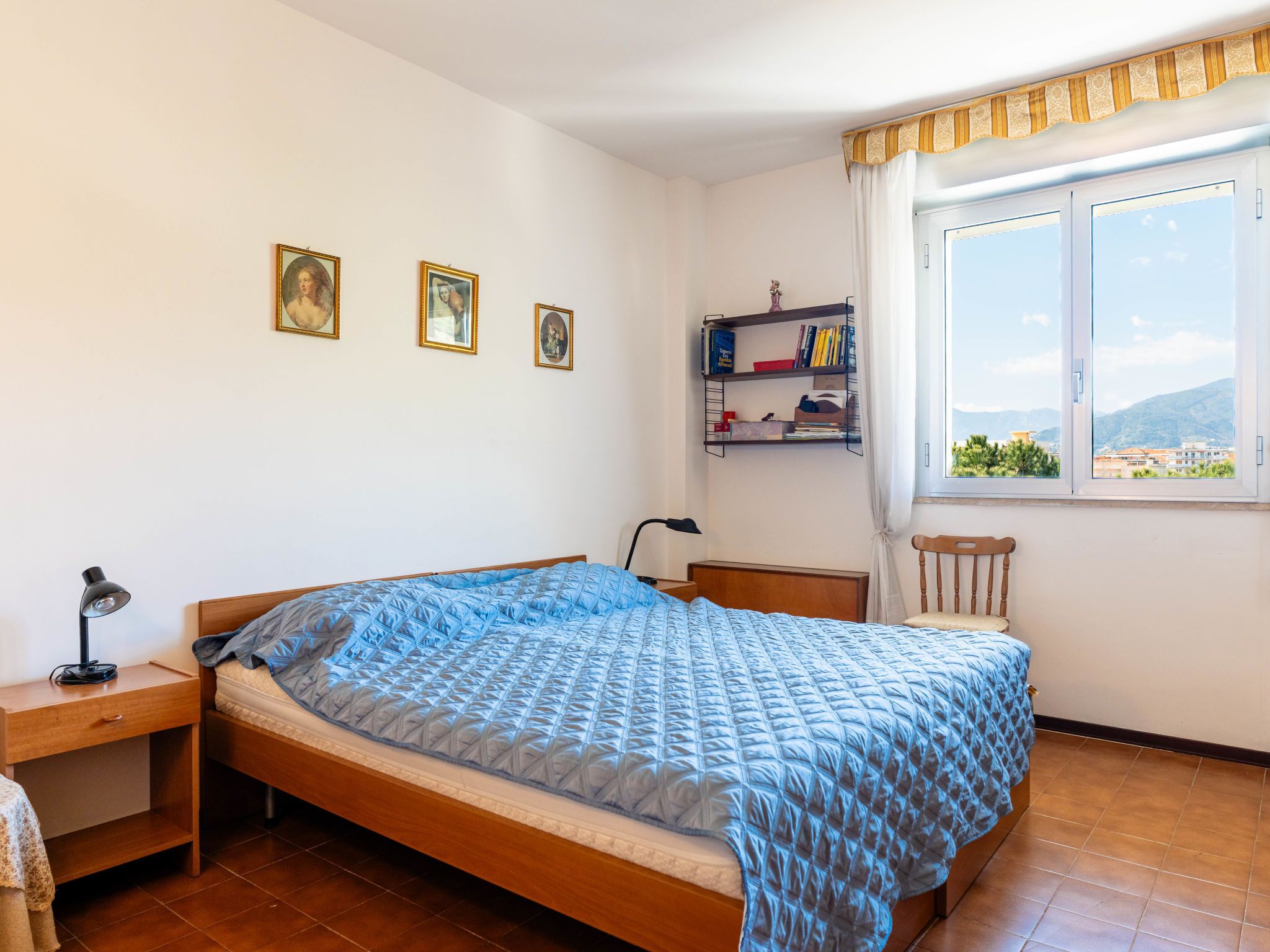 Un dormitorio con cama, escritorio, silla, ventana con vistas y estante con libros y objetos decorativos.