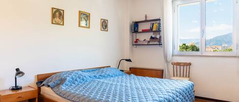 Un dormitorio con cama, escritorio, silla, ventana con vistas y estante con libros y objetos decorativos.