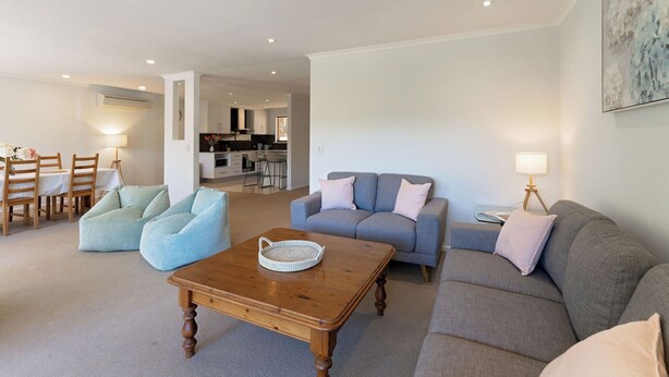 A modern living room with a grey sofa, a blue armchair, and a wooden coffee table. There is a dining area with wooden chairs and a table, and a kitchen in the background.