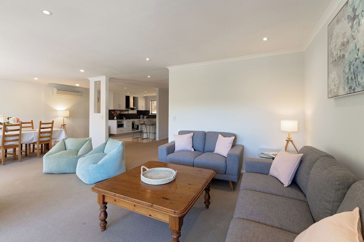 A modern living room with a grey sofa, a blue armchair, and a wooden coffee table. There is a dining area with wooden chairs and a table, and a kitchen in the background.