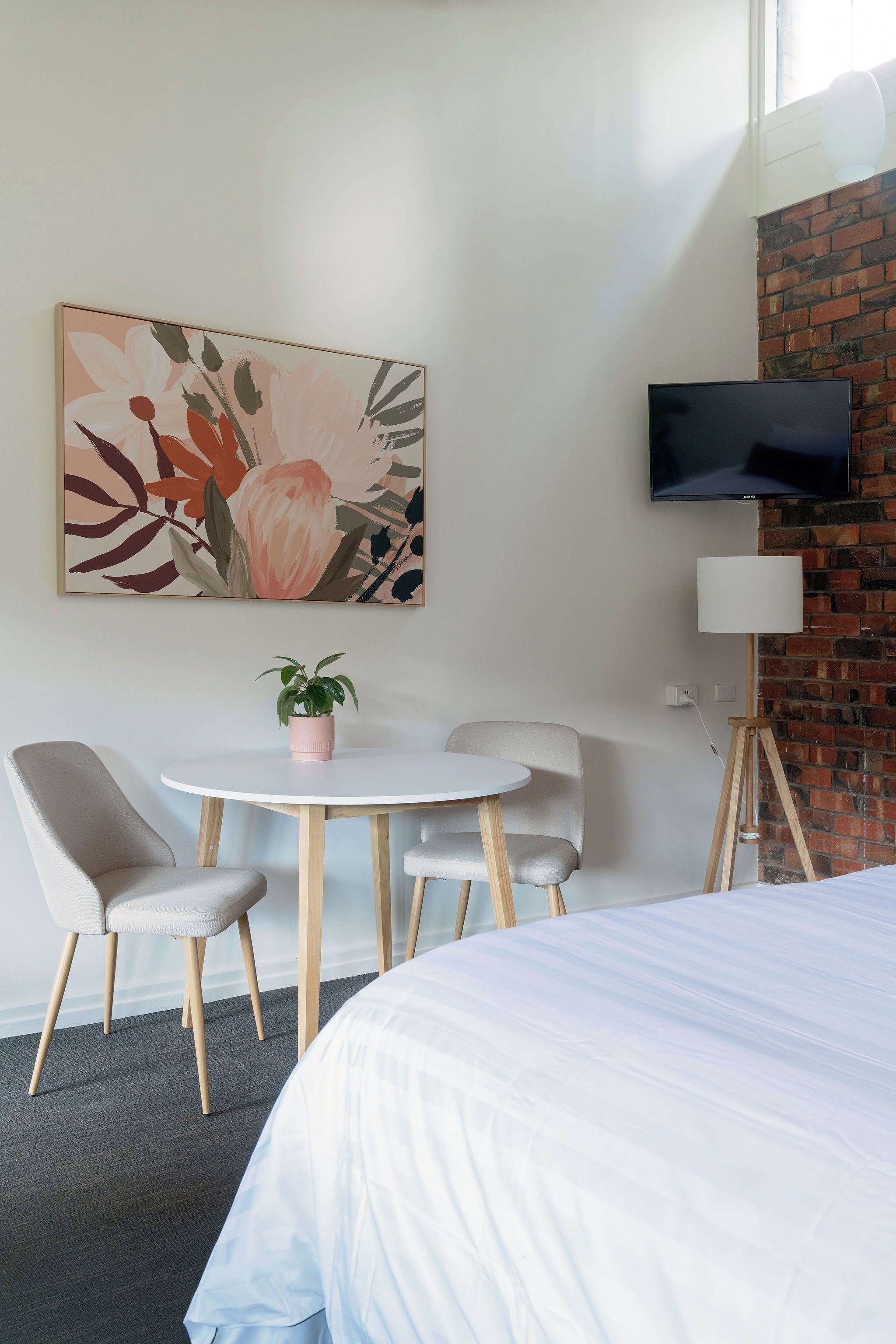 A hotel room with a bed, a round table with chairs, a potted plant, a flat-screen TV, and a painting on the wall.
