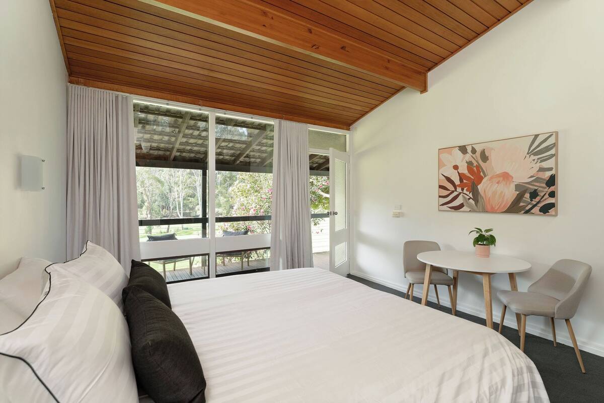 A bedroom with a large bed, a dining table with chairs, a potted plant, and a painting on the wall.