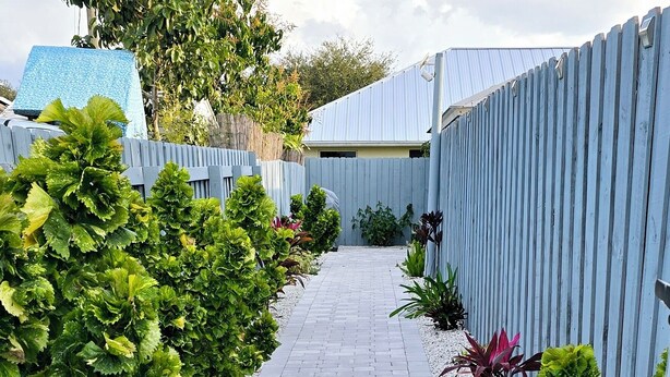 Un sendero conduce a una casa con felpudo que dice 'WELCOME', rodeada de setos podados y una cerca azul.