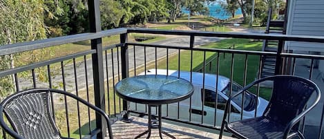 A balcony with two wicker chairs, a small round table, and a view of a white vehicle and greenery.