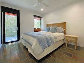 Une chambre à coucher avec un lit, un ventilateur de plafond, une fenêtre donnant sur un espace verdoyant et une porte-fenêtre donnant sur un balcon.