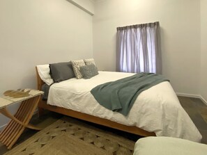 Un letto rifatto con coperta bianca e copriletto verde, un comodino in legno con una lampada, e un tappeto decorato su un pavimento in legno.
