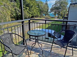 Een balkon met twee rieten stoelen, een klein rond tafeltje en uitzicht op een wit voertuig en groen.
