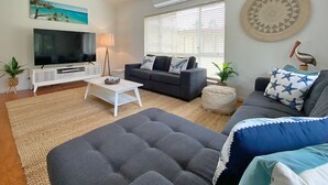 A living room with a flat-screen TV, a grey sofa, a white coffee table, and a large window with blinds.