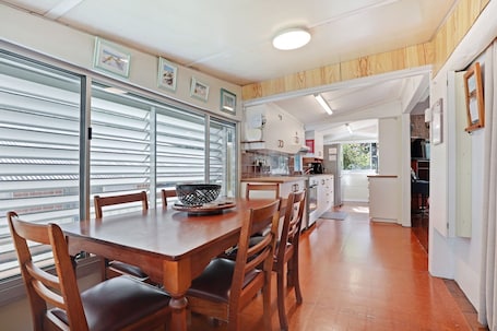 A dining area with a wooden table and chairs, a large window with blinds, and a view of the outside.. Original house, large gardens in bush setting