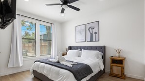 Un dormitorio con una cama, un ventilador de techo, una ventana con persianas y dos cuadros en la pared.