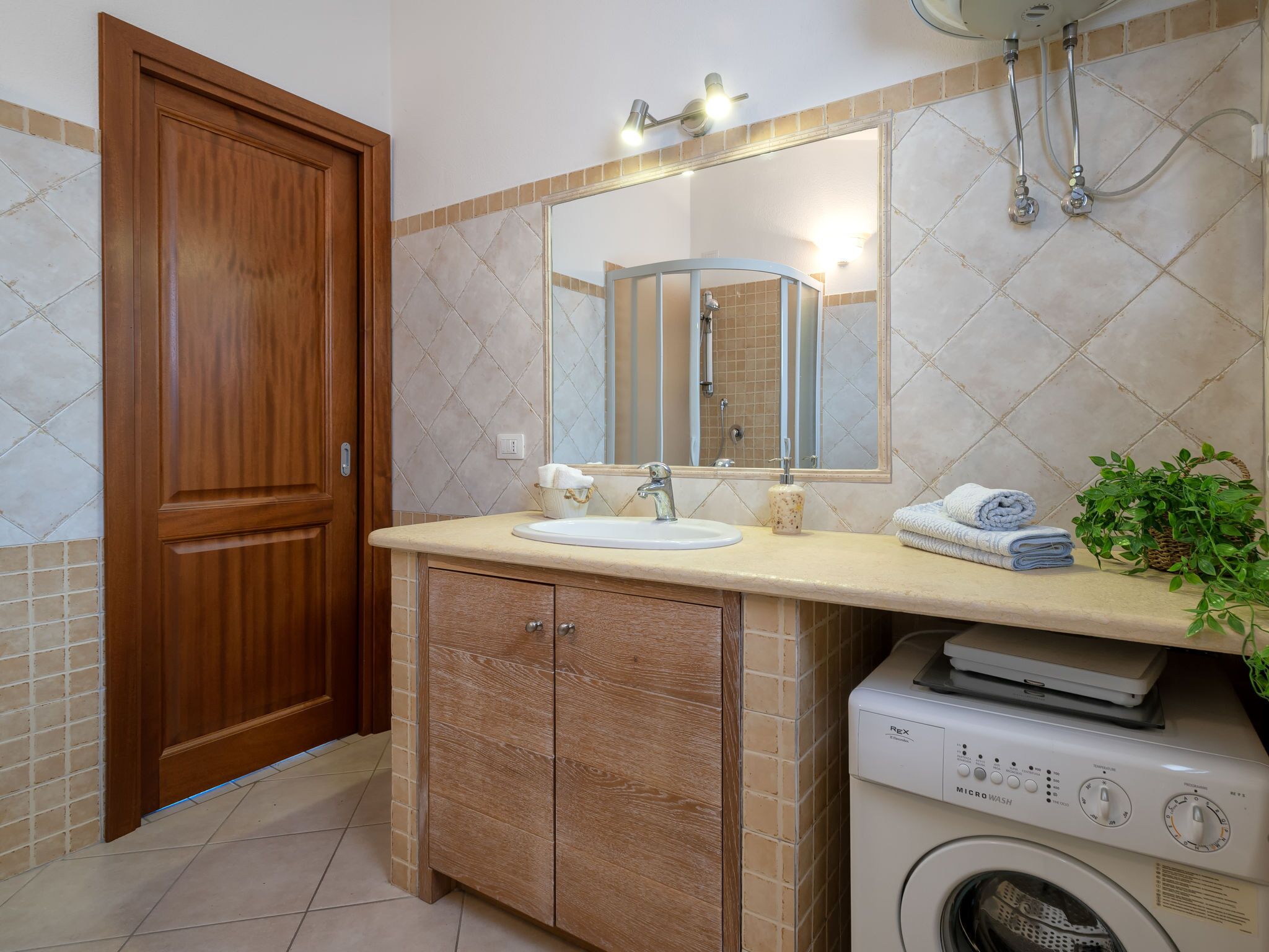 A bathroom with a wooden door, a sink with a mirror, a washing machine, and a potted plant.