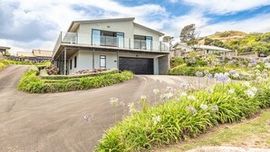 A modern two-story house with a garage, surrounded by well-maintained gardens and a driveway. - Sunset Escape - Stunning Beachfront Views (Kai Iwi)