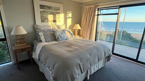 A bedroom with a large bed, two bedside tables, a window with a view of the ocean, and a ceiling light. - Sunset Escape - Stunning Beachfront Views (Kai Iwi)