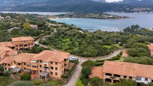 A coastal residential area with terracotta-roofed buildings, lush greenery, and a view of the sea and mountains in the background.
