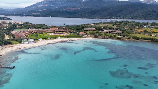 An aerial view of a coastal resort with a beachfront, villas, and a mountainous backdrop.