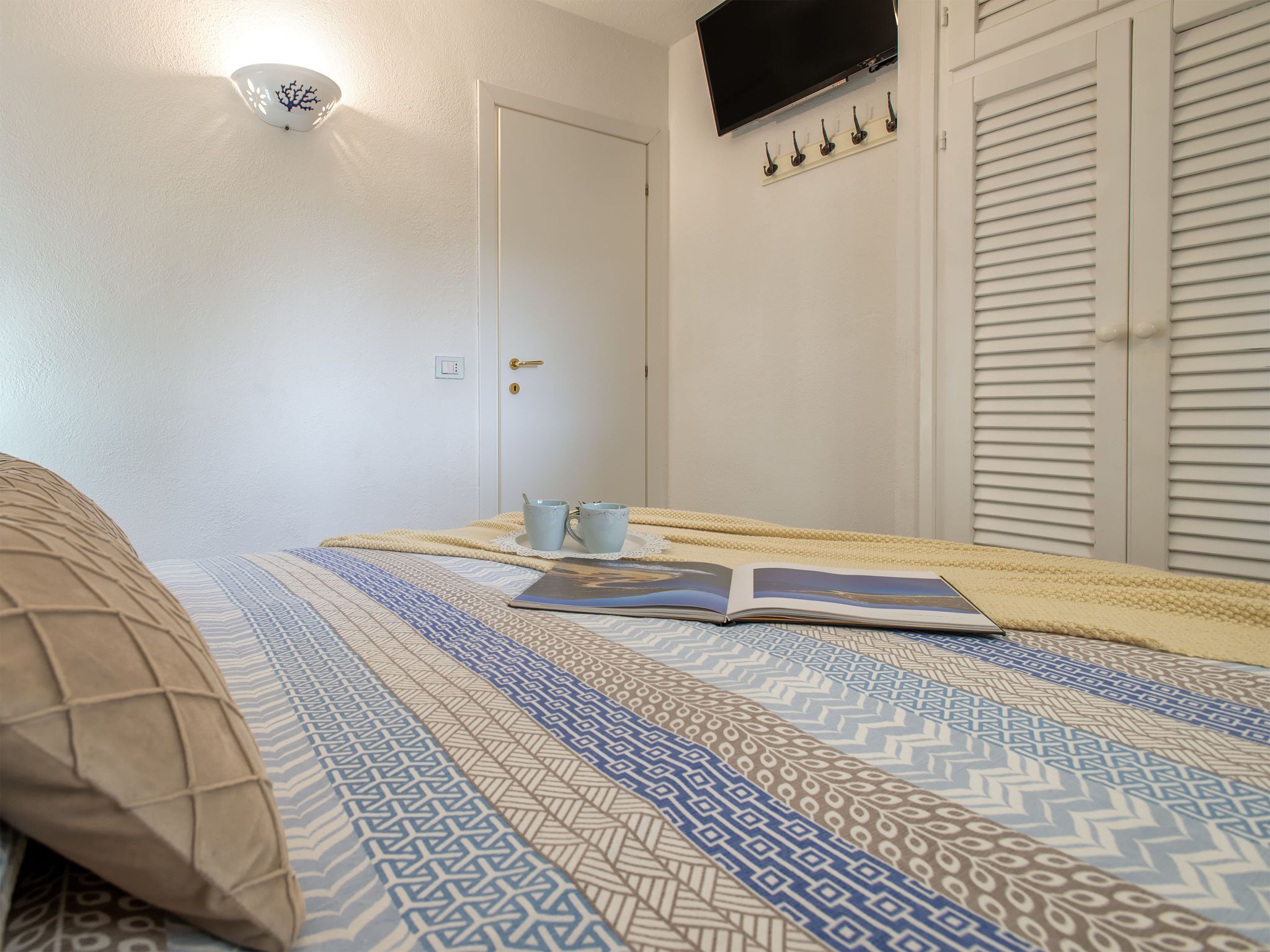 A bedroom with a bed, a bedside table with two cups, a book, a wall-mounted TV, and a coat rack.