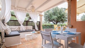 An outdoor patio with a dining table set for two, a sofa, and a small table with a vase of flowers.