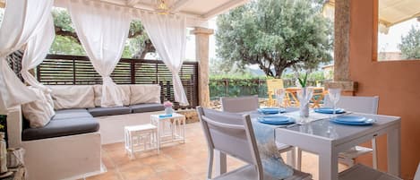 An outdoor patio with a dining table set for two, a sofa, and a small table with a vase of flowers.