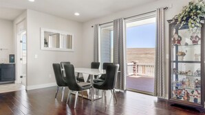 Un comedor con una mesa blanca y sillas negras, un aparador de vidrio con objetos decorativos y vista a la playa a través de puertas abiertas.