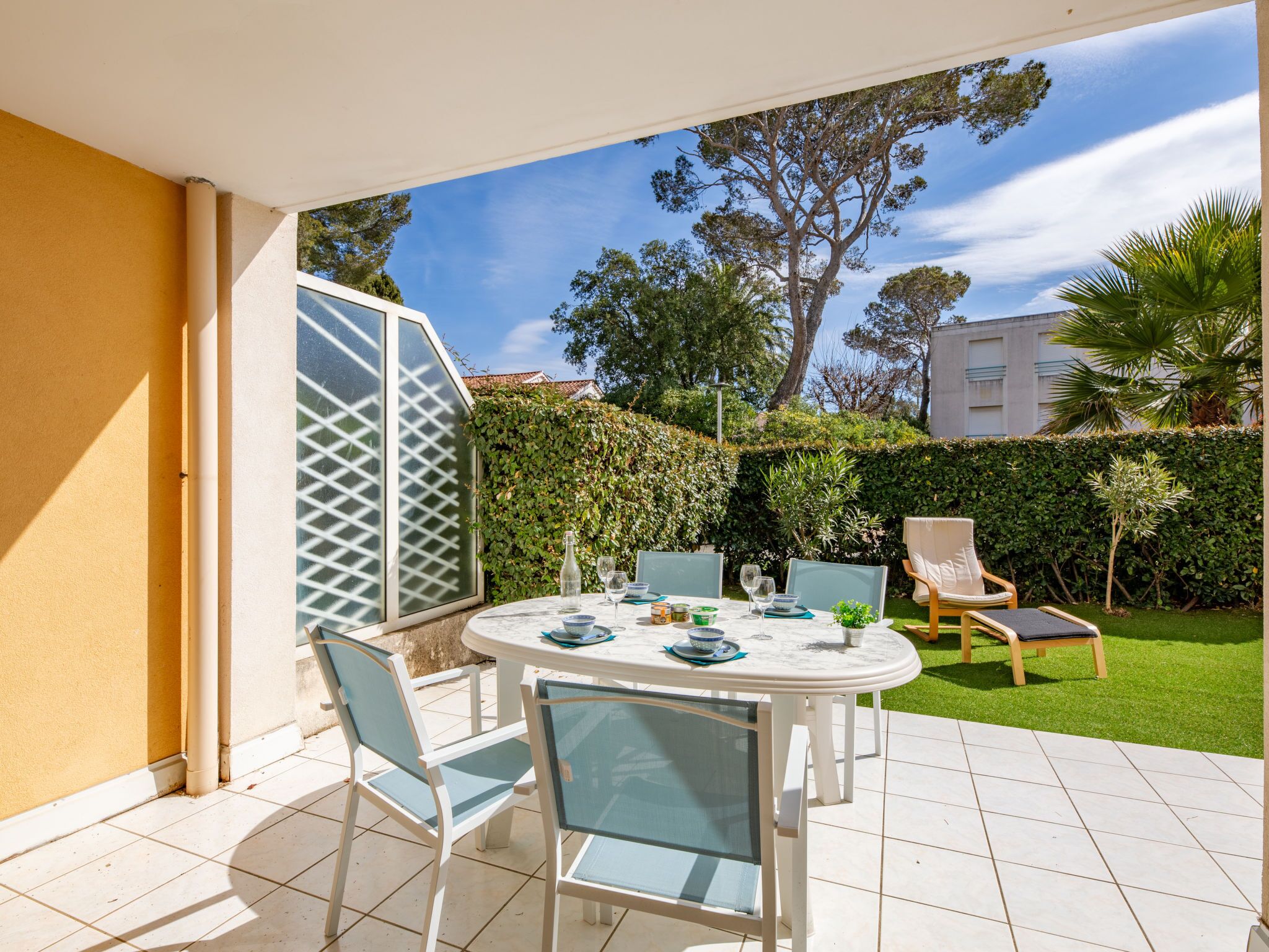 Pátio com mesa redonda e cadeiras, porta de vidro e vista para um jardim e um prédio.