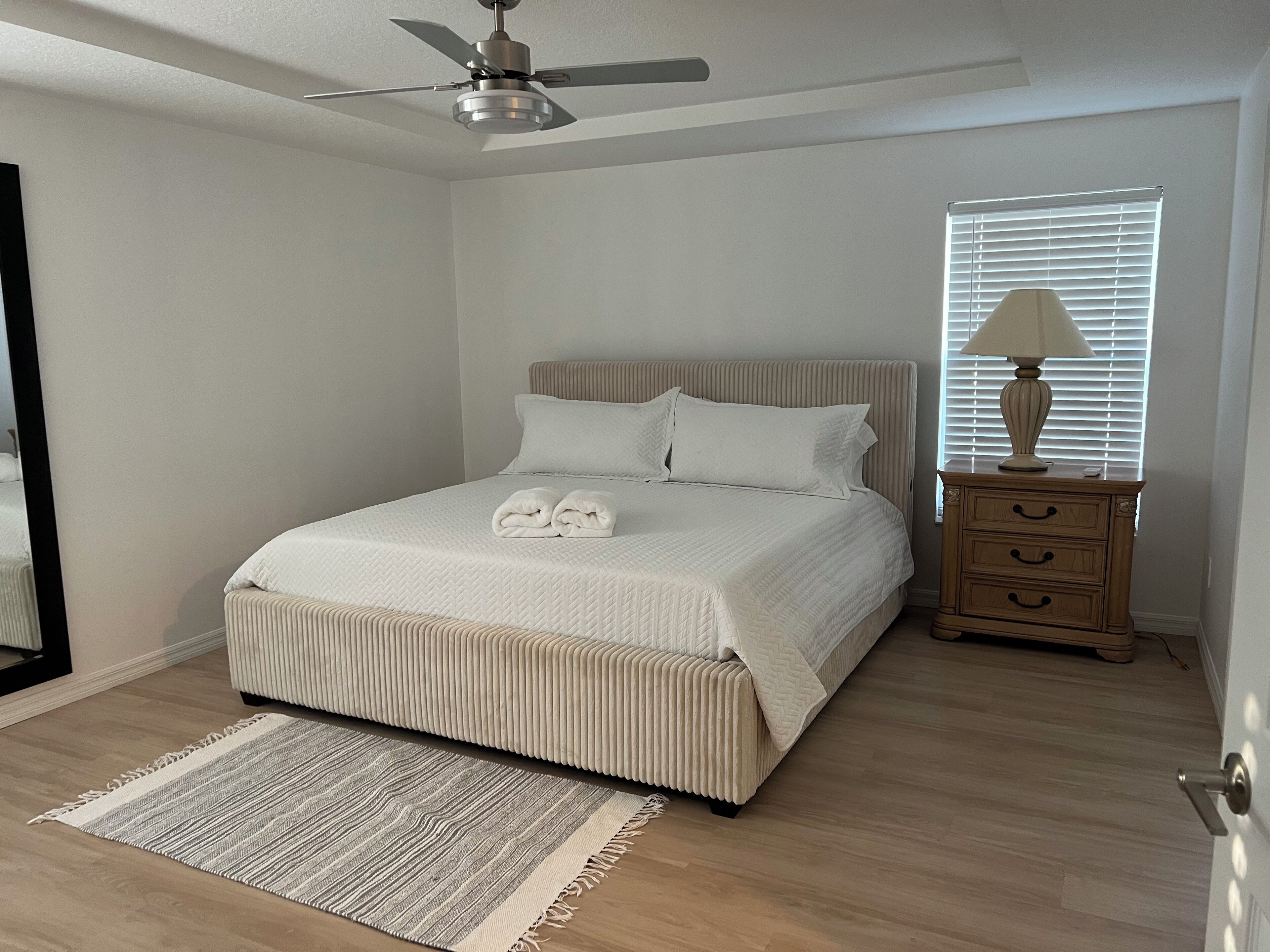 Quarto com cama, mesa de cabeceira com abajur, ventoinha no teto e janela com persianas.