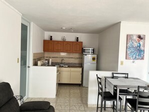 A compact kitchen with wooden cabinets, a microwave, and a refrigerator. There is a dining table with chairs and a small sofa in the foreground.