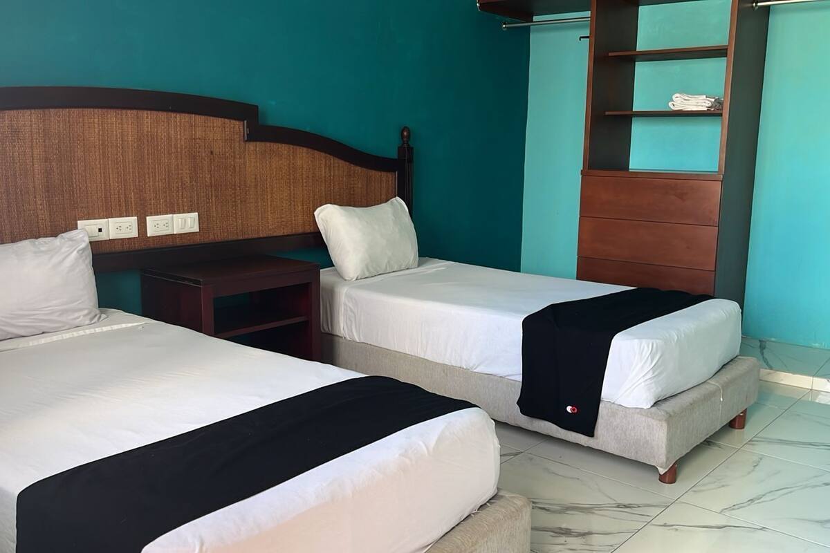 A hotel room with two beds, a wooden headboard, a small wooden nightstand, and a shelf unit against the wall.