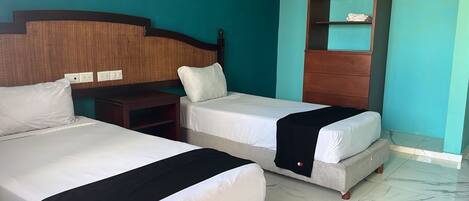 A hotel room with two beds, a wooden headboard, a small wooden nightstand, and a shelf unit against the wall.