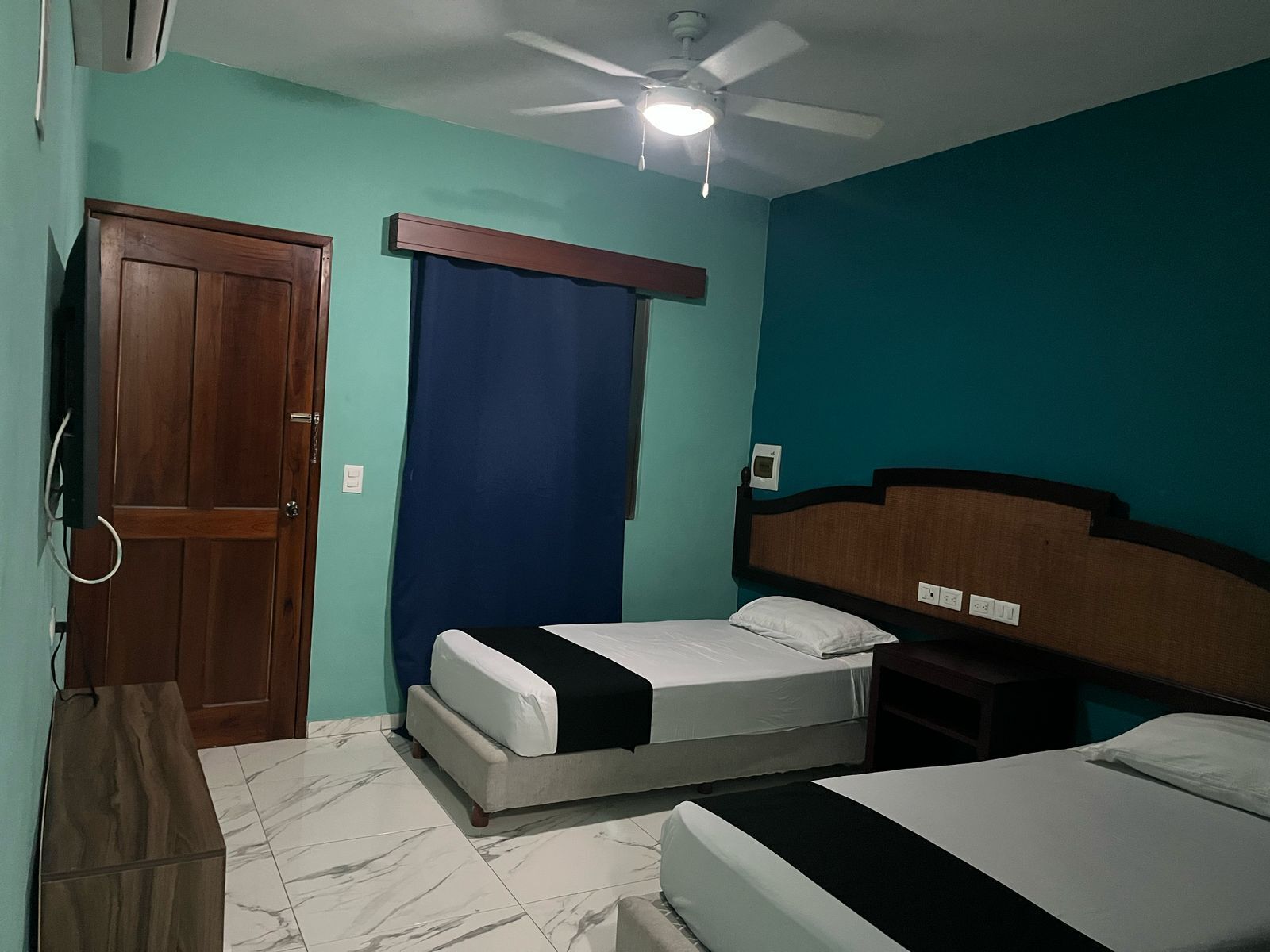 A room with two beds, a ceiling fan, a wooden door, and a blue curtain.
