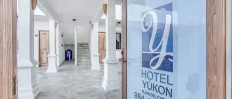 Entrada de hotel con piso de mármol, pilares blancos y puerta de cristal que muestra el nombre y número de contacto del hotel.