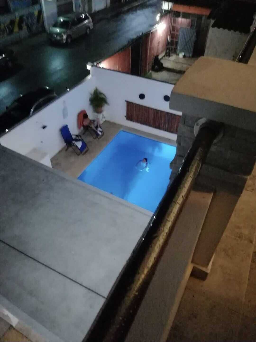 Una piscina en la azotea con agua azul, una pared blanca y una cerca de madera. Hay dos sillas reclinables azules y una planta en maceta.