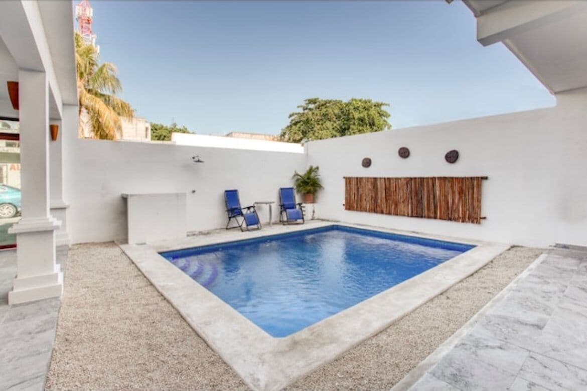 A swimming pool with two lounge chairs, a white wall with a wooden panel, and a clear sky.