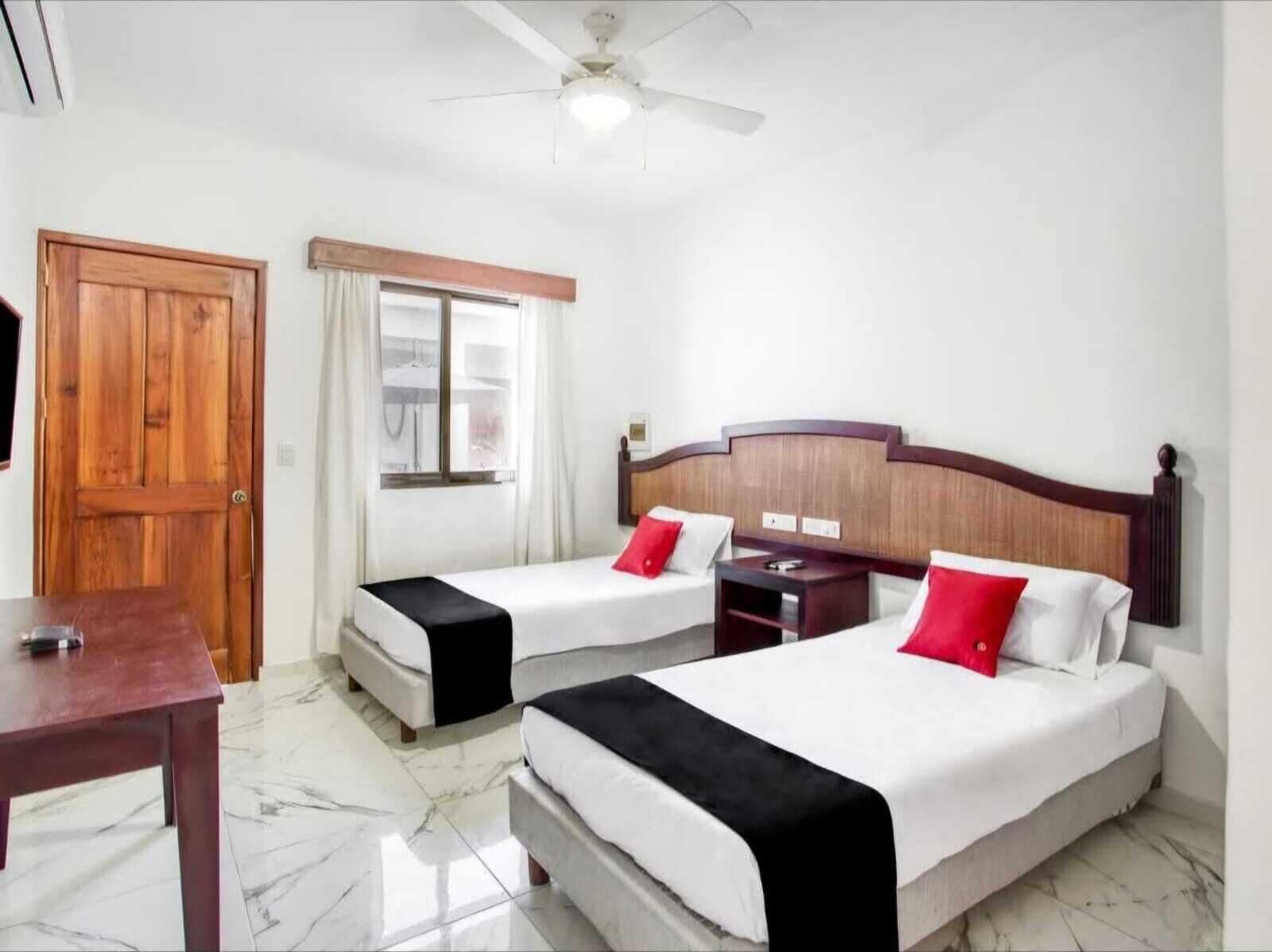 A hotel room with two beds, a wooden door, a window with curtains, a ceiling fan, and a marble floor.