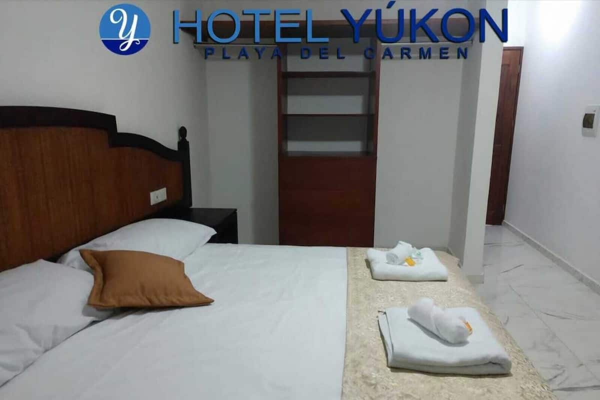 A hotel room with a bed, a nightstand, a wooden headboard, a brown pillow, and a folded towel on a beige patterned table.