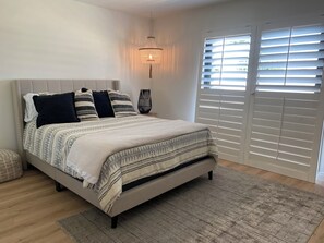 Ein Schlafzimmer mit Bett, Nachttisch, Lampe und einem Fenster mit Jalousien.