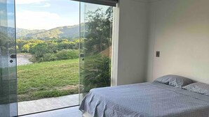 Une chambre dotée d’une grande porte-fenêtre coulissante donnant sur un balcon, d’un lit avec une literie grise et offrant une vue sur un paysage verdoyant.