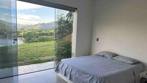Un dormitorio con un amplio ventanal corredera que da a un balcón con vistas a un paisaje verde y colinas.