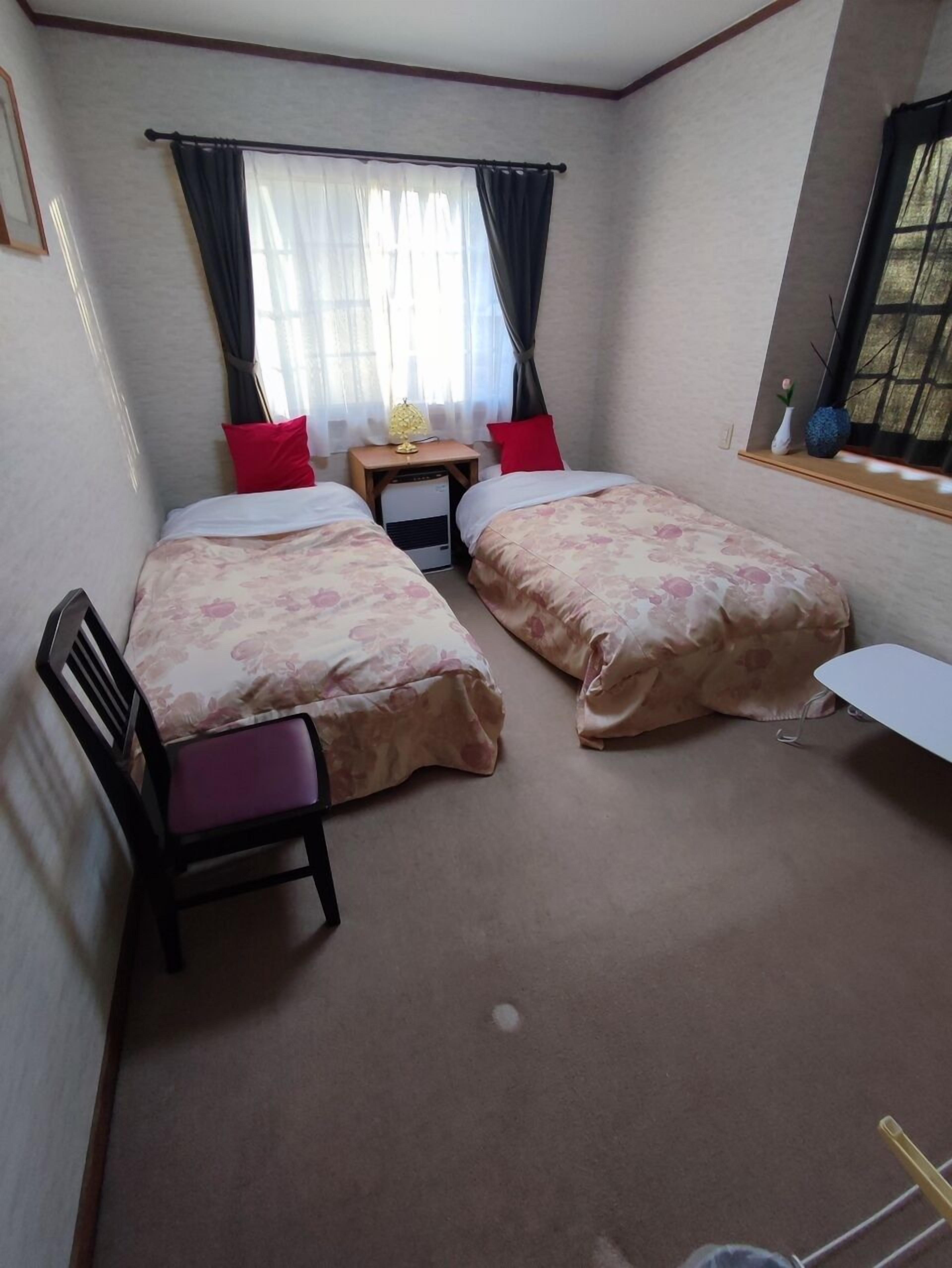 Una habitación pequeña y limpia con dos camas, un escritorio, una silla, un ventanal con cortinas y una mesita con lámpara.