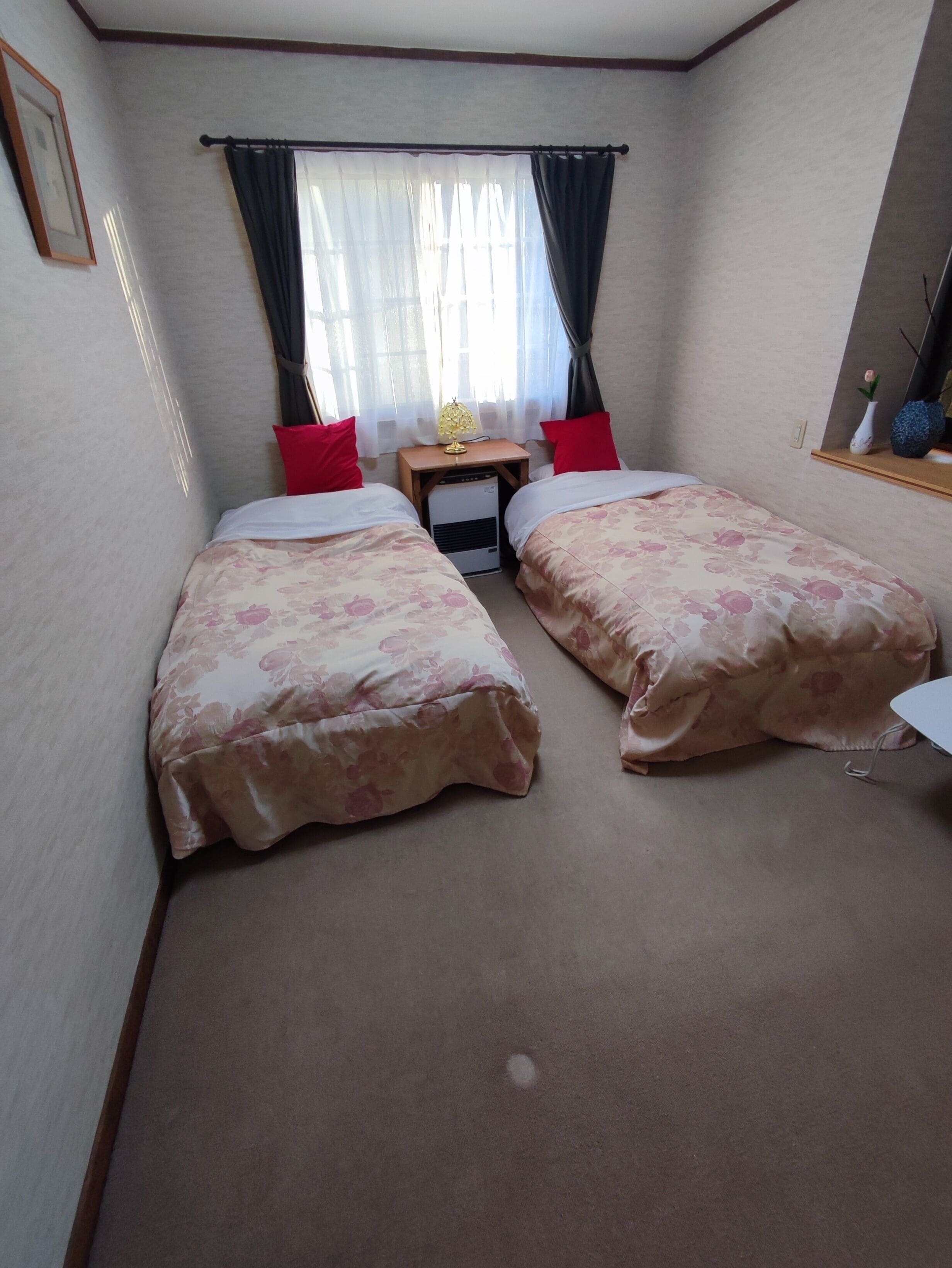A small, neatly arranged bedroom with two beds, a bedside table with a lamp, and a small heater.