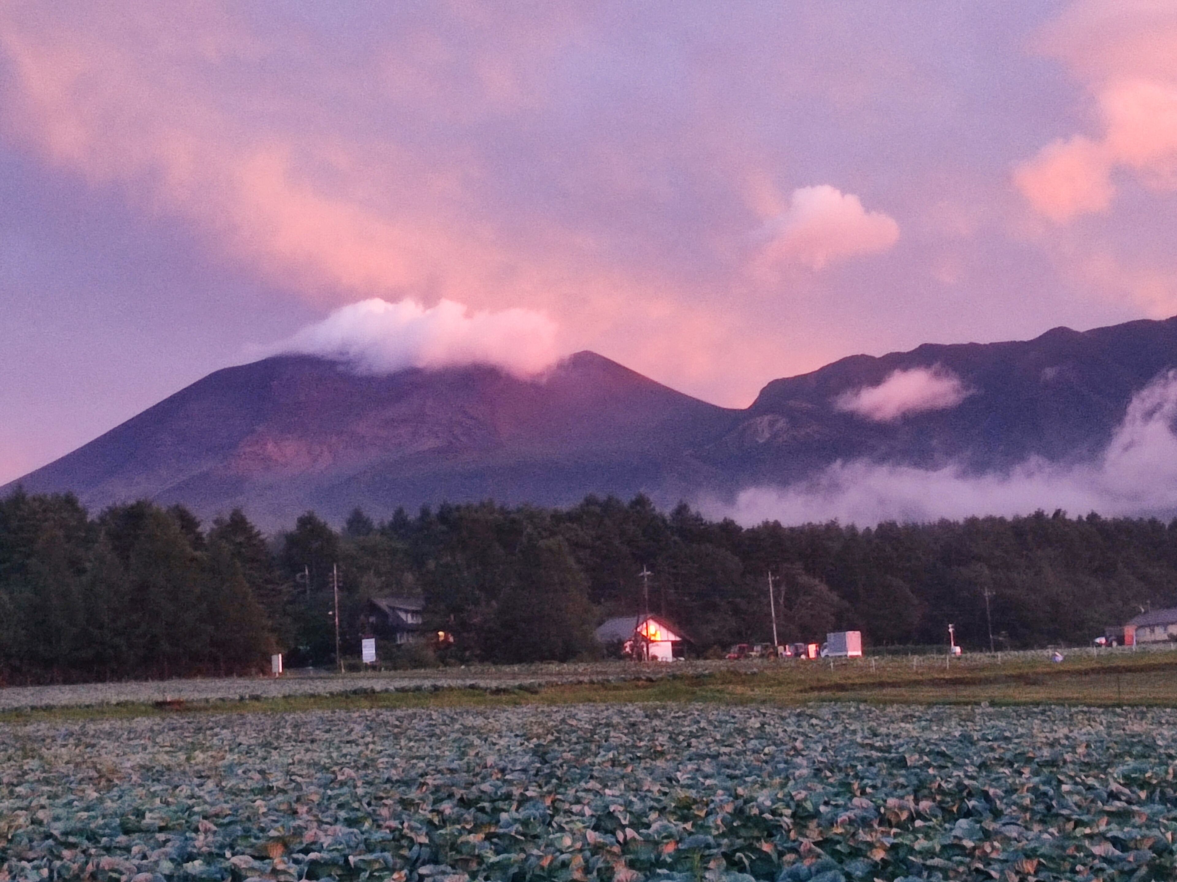 A mountain landscape with a field of crops in the foreground and a small building with a red roof in the middle ground.