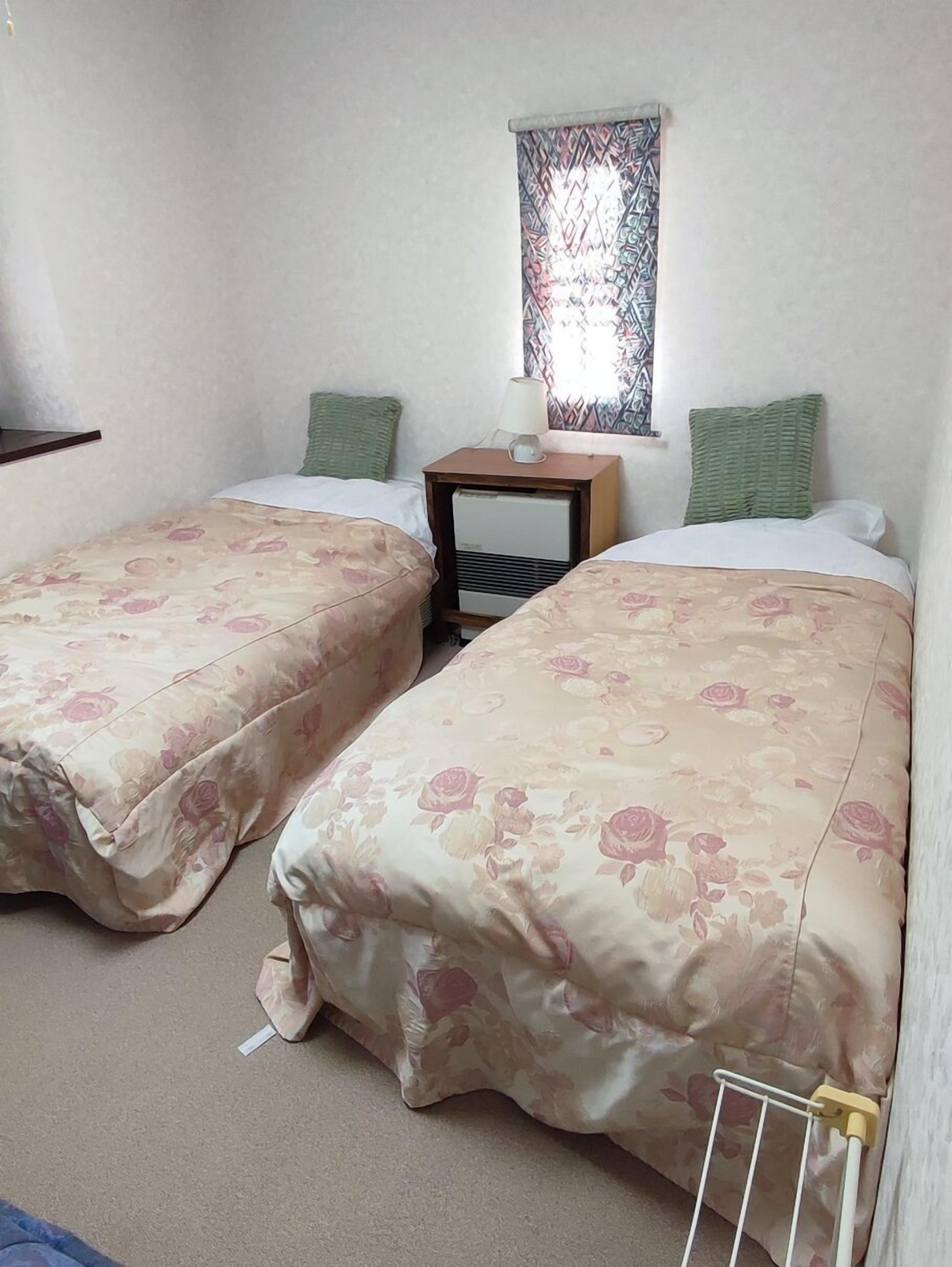 Una habitación con dos camas, una mesita de noche, una lámpara y una ventana con cortina estampada.