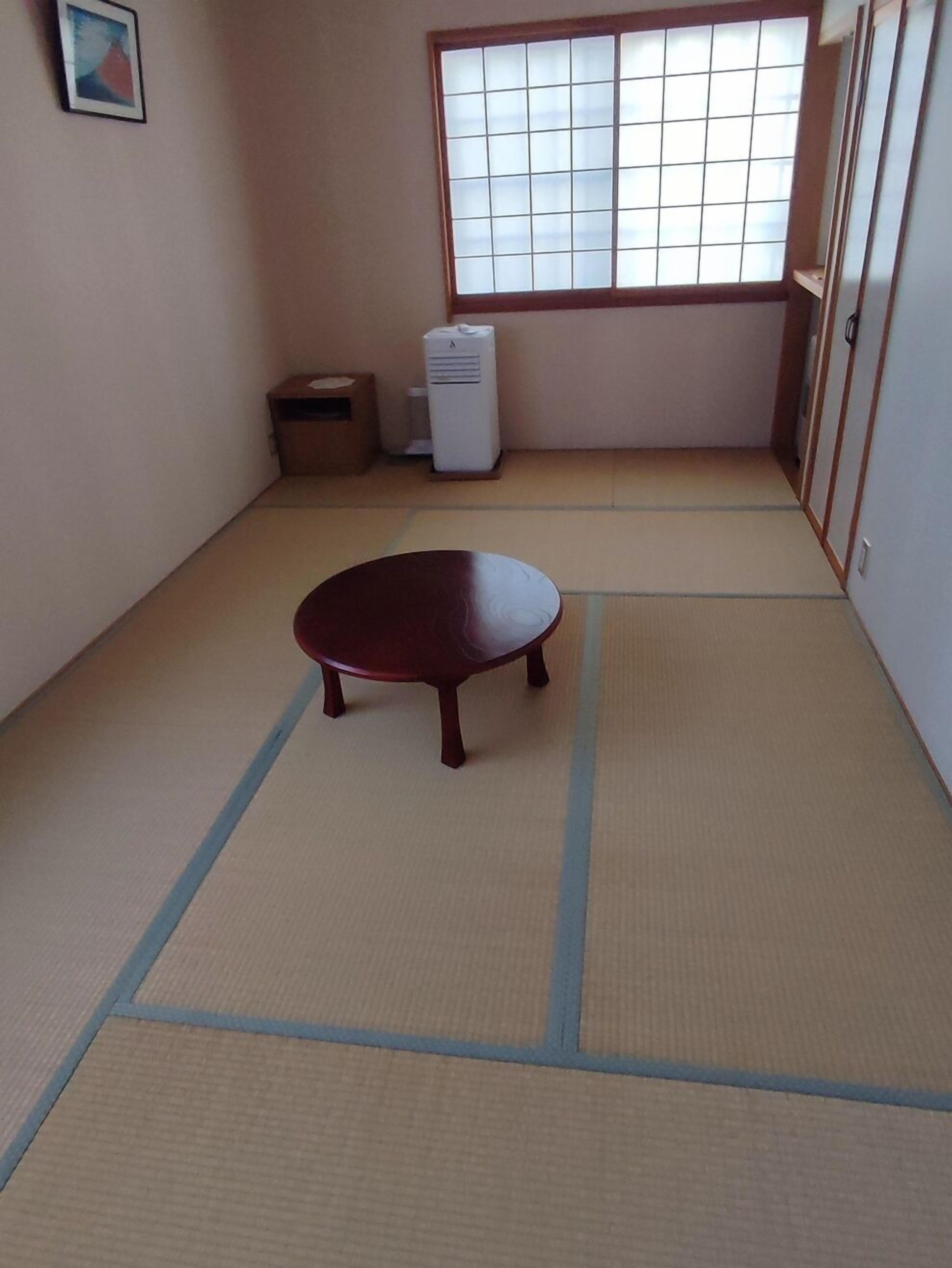 Una habitación con suelo de tatami, una mesa de tatami, un futón, una ventana con diseño de cuadrícula y un aire acondicionado blanco.