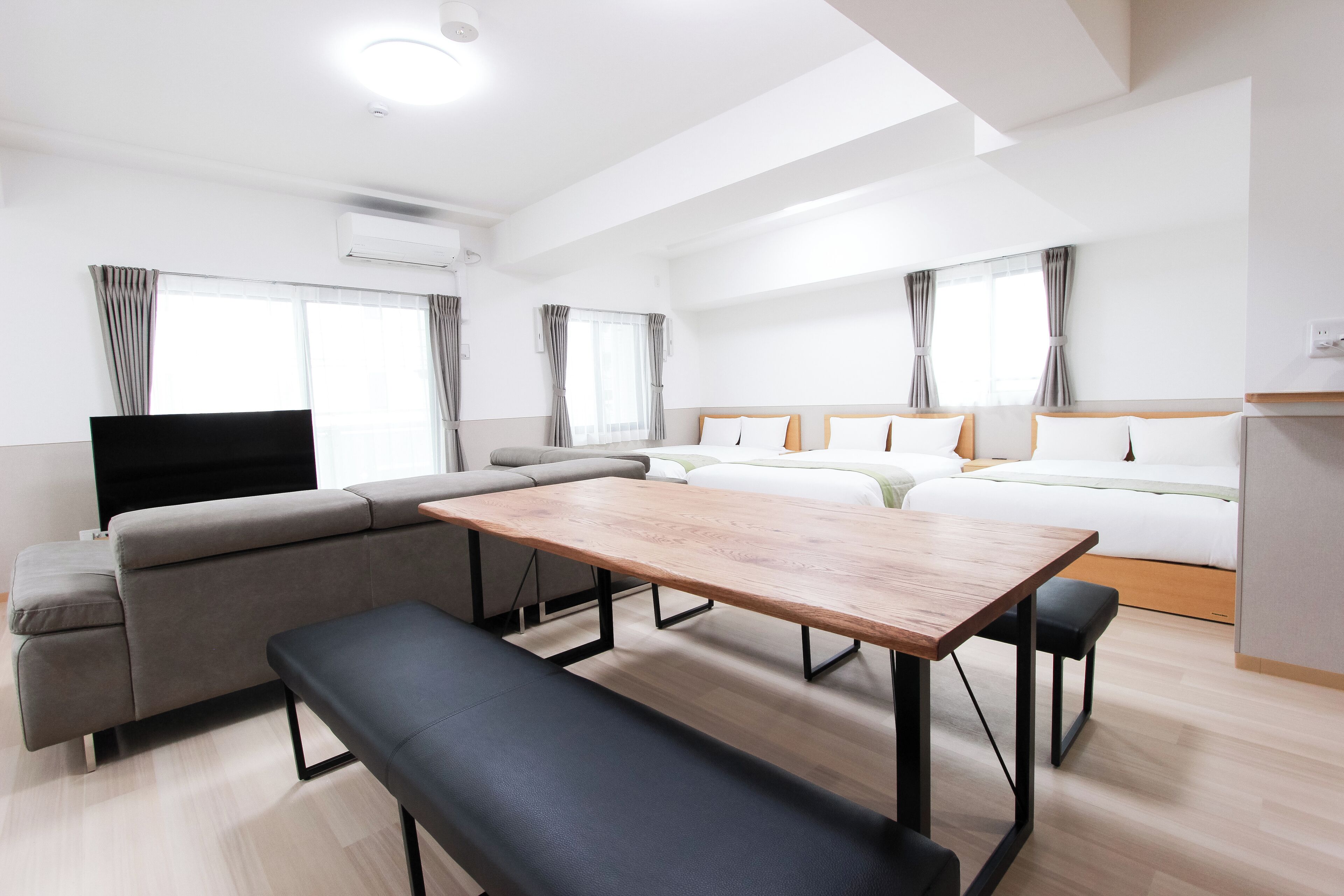 A modern hotel room with a large wooden dining table, grey sofa, and a flat-screen TV.