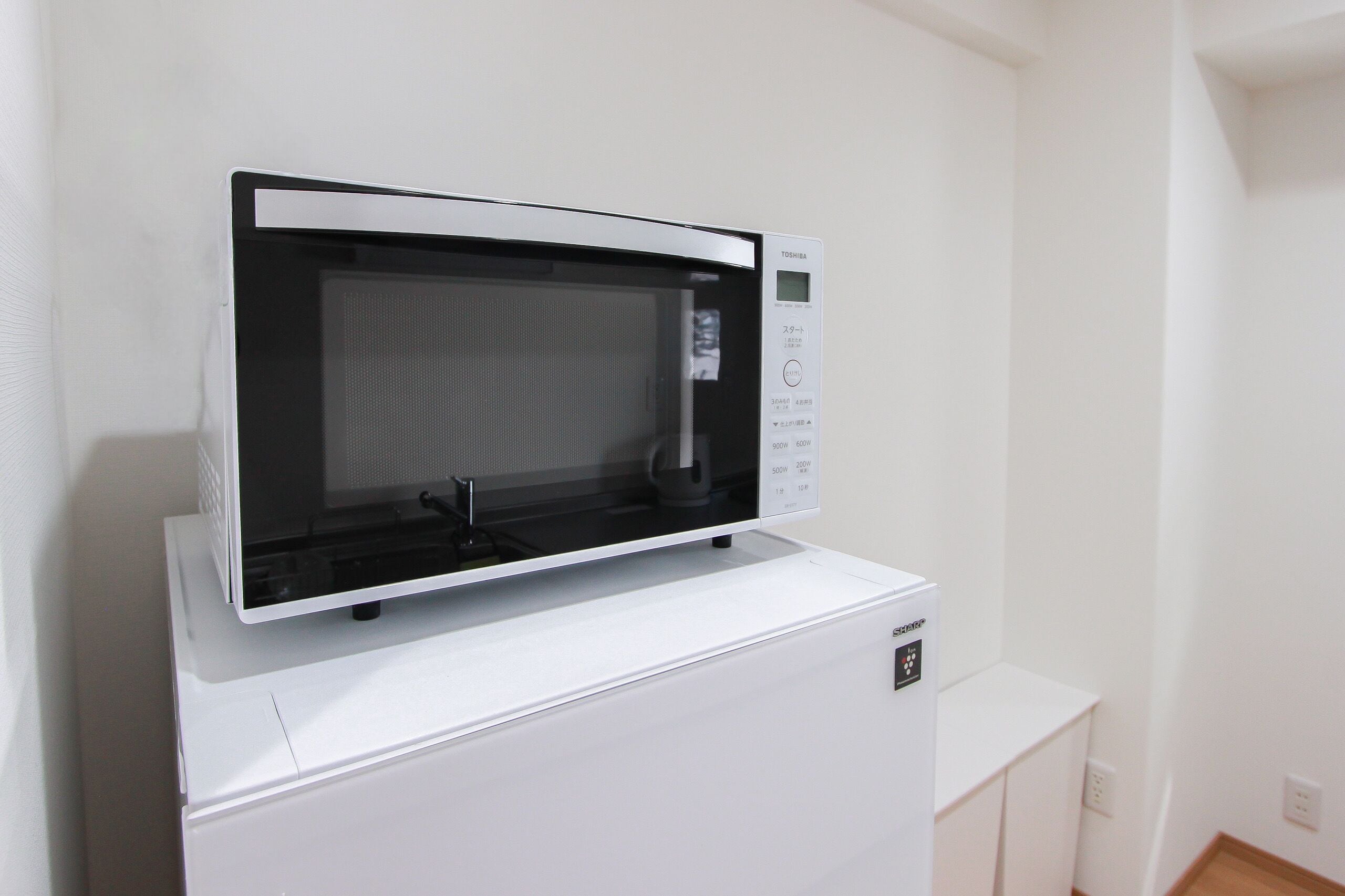 A compact microwave oven with a digital display and control panel, placed on top of a white appliance, likely a refrigerator, in a clean and minimalistic room.