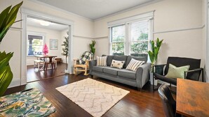 A living room with a grey sofa, a wooden coffee table, and a patterned rug. There are two chairs, a plant, and a window with blinds. - 240- History Buff, Sleeps 10, Central Location to FUN, Pet & Family Friendly (Jacksonville)