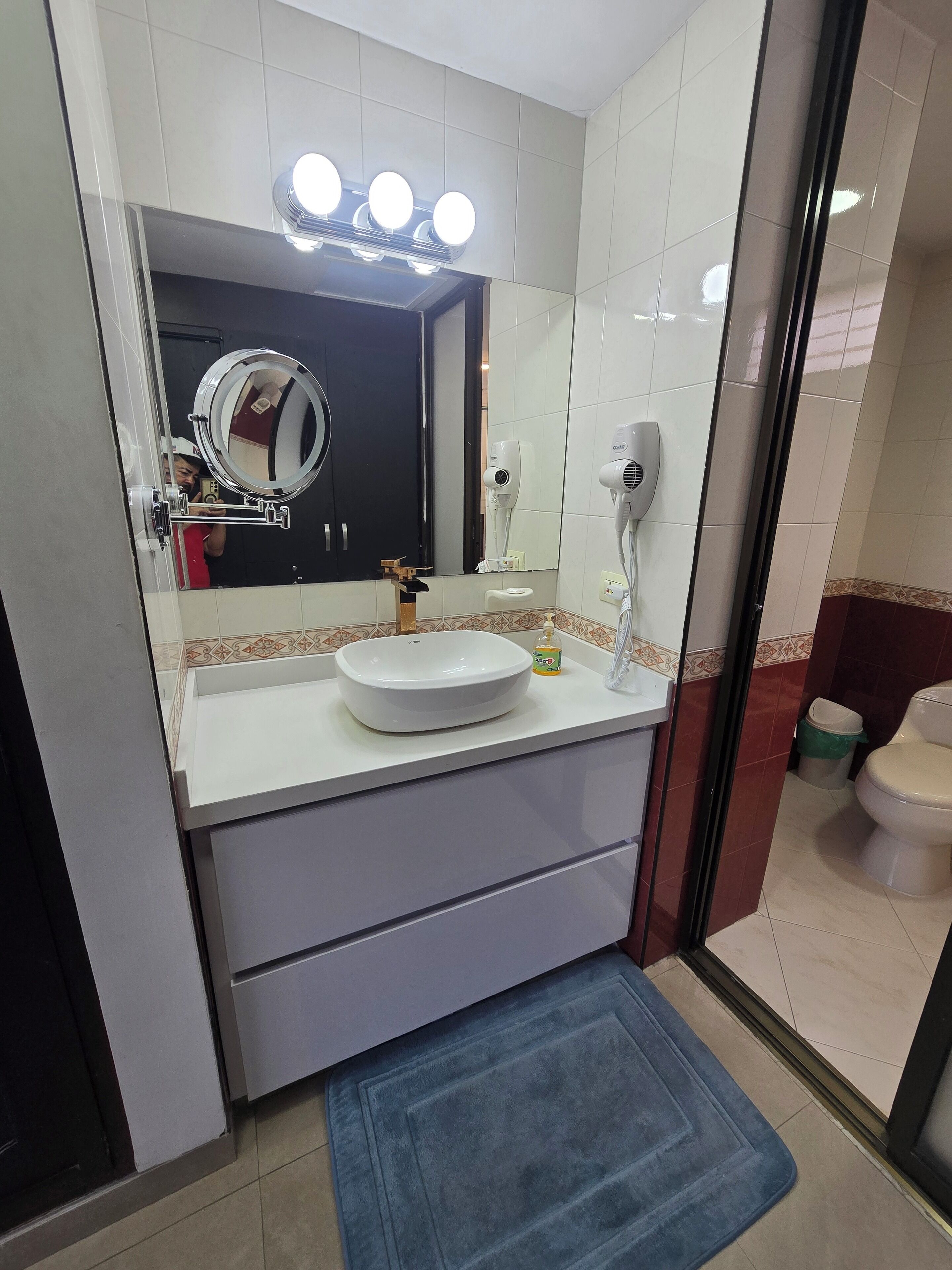 Un baño con lavamanos blanco, un espejo con luces, un secador de pelo y un tapete azul.
