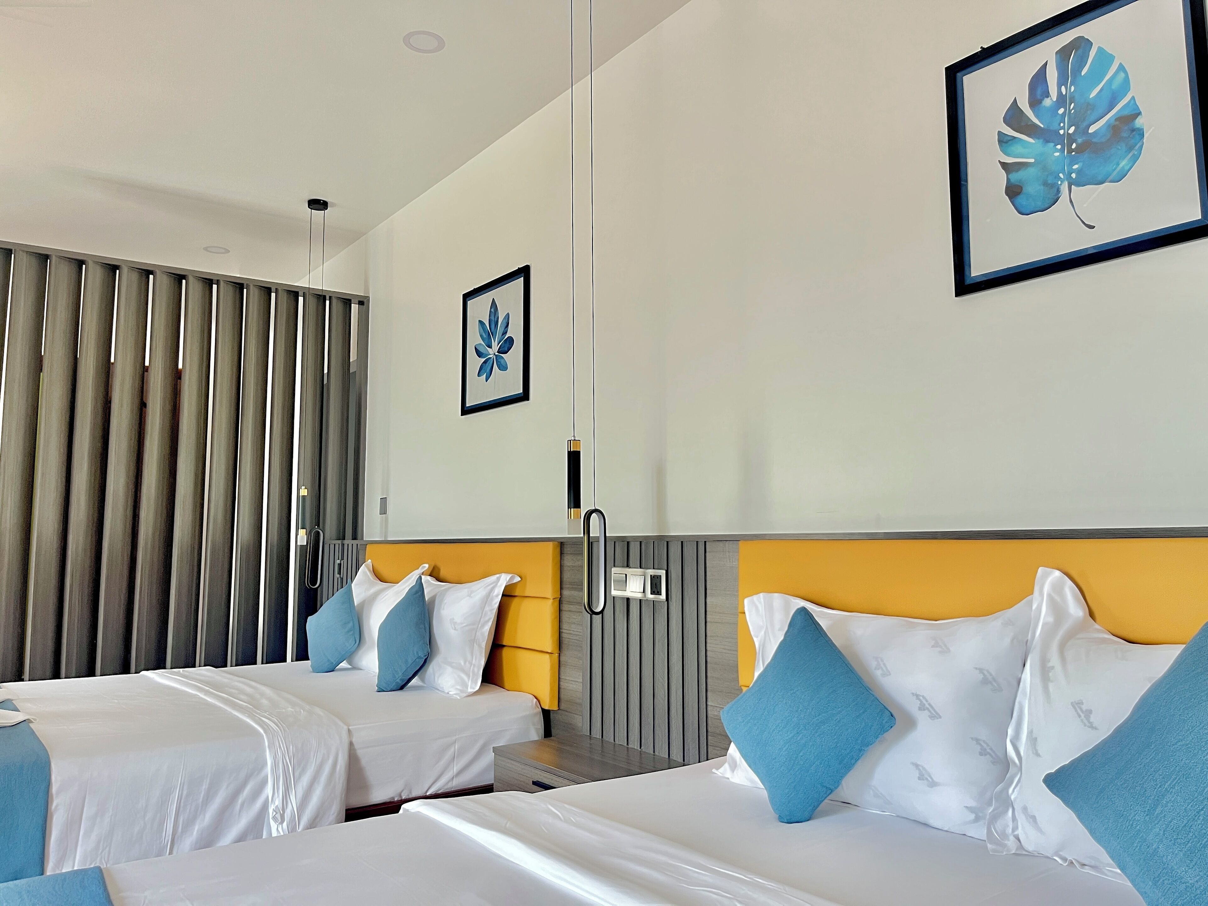 A hotel room with two beds, each with white bedding and blue pillows, a yellow headboard, and a framed blue leaf artwork on the wall.