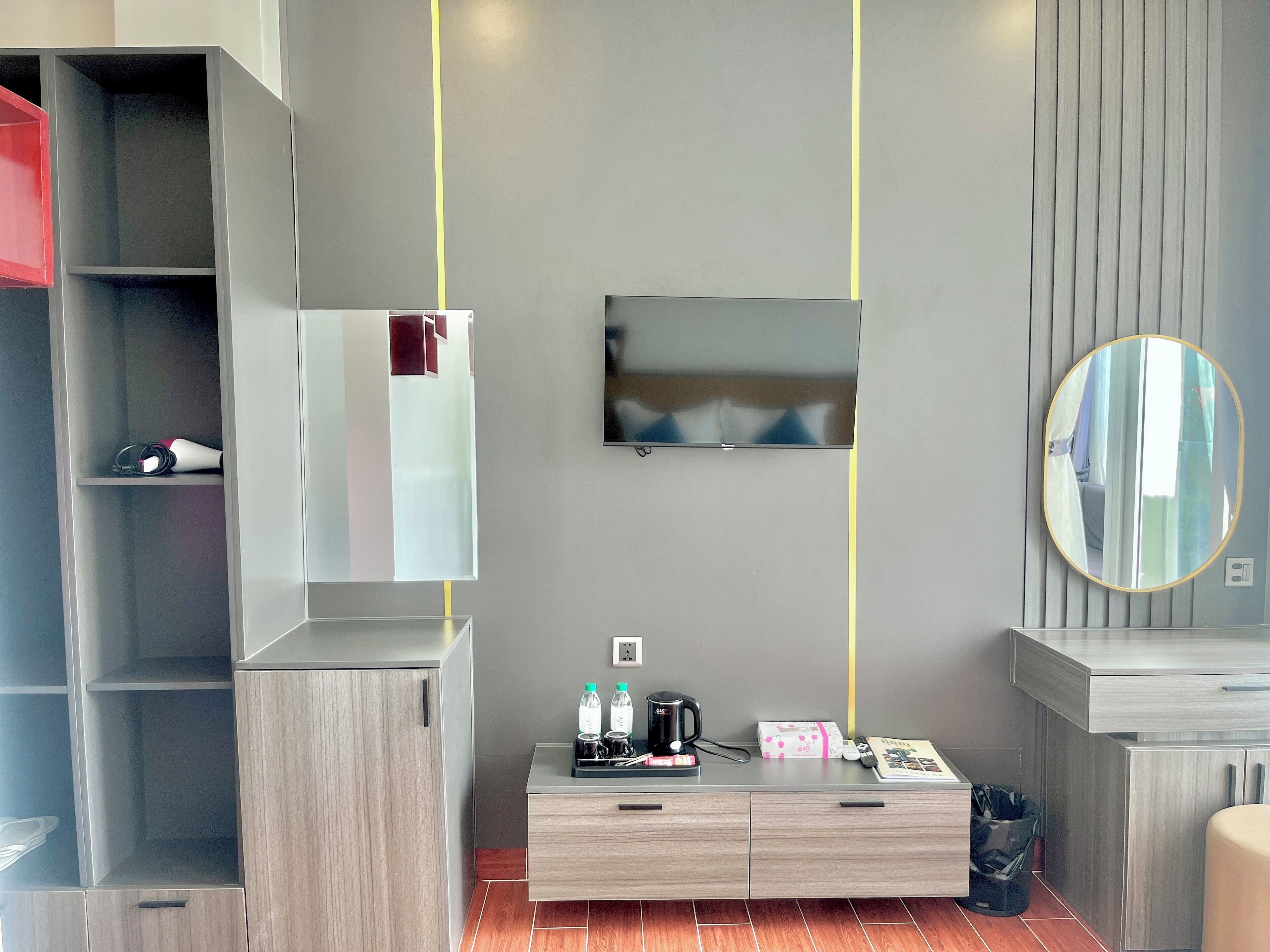 Habitación de hotel moderna con una pared gris, un televisor de pantalla plana sobre un tocador de madera, un espejo y piso de madera.