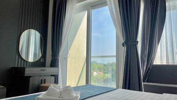 A hotel room with a bed, a blue bedspread, a set of white towels, a round mirror, and a view of greenery and buildings through a large window. - Kep Sea View Hotel (Kep)